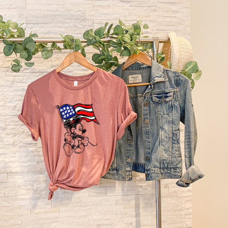 Puede incluir: Una camiseta rosa con una imagen de dibujos animados de Mickey Mouse sosteniendo una bandera estadounidense. La camiseta est&aacute; colgada en una percha con una chaqueta vaquera.