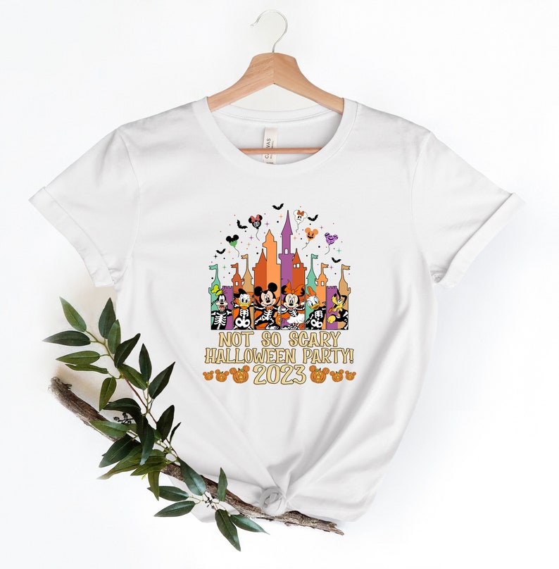 May include: White t-shirt featuring a colourful graphic of Mickey Mouse and his friends in front of a castle. The text "Not So Scary Halloween Party! 2023" is printed on the shirt.