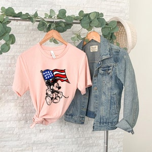 Puede incluir: Una camiseta rosa con una imagen de dibujos animados de Mickey Mouse sosteniendo una bandera estadounidense. La camiseta est&aacute; en una percha con una chaqueta vaquera colgada a su lado.