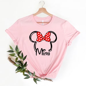 Op de afbeelding: Roze T-shirt met een zwarte en witte omtrek van Mickey Mouse oren met een rode strik met witte stippen. De tekst "Mimi" is onder de oren gedrukt.