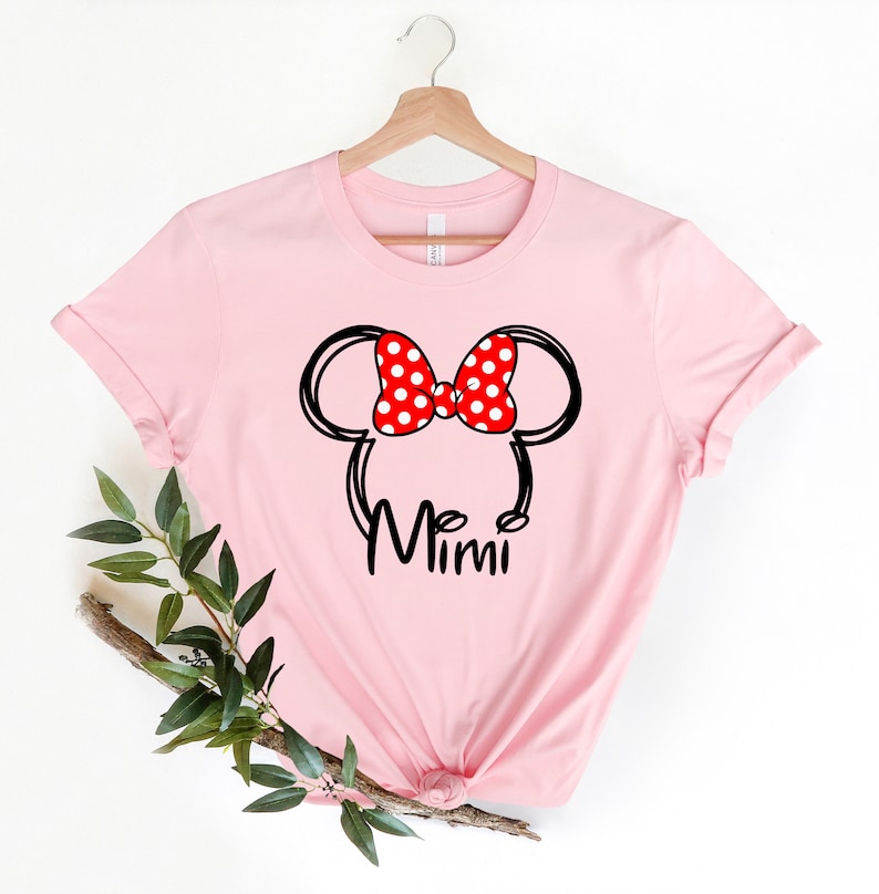Op de afbeelding: Roze T-shirt met een zwarte en witte omtrek van Mickey Mouse oren met een rode strik met witte stippen. De tekst "Mimi" is onder de oren gedrukt.
