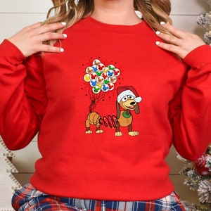 Pu&ograve; includere: Una felpa rossa con un bassotto dei cartoni animati con un cappello da Babbo Natale. Il bassotto tiene in mano dei palloncini con le facce di Topolino. La felpa &egrave; un design festivo natalizio.