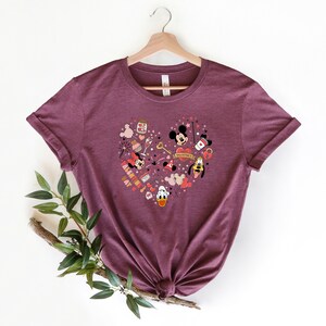 Pode incluir: Uma camiseta vinho com um design em forma de cora&ccedil;&atilde;o que apresenta Mickey Mouse, Minnie Mouse, Donald Duck e Pateta. O design inclui v&aacute;rios elementos tem&aacute;ticos do Dia dos Namorados como cora&ccedil;&otilde;es, estrelas e uma chave. O texto "Valentine's Day" est&aacute; inclu&iacute;do no design.