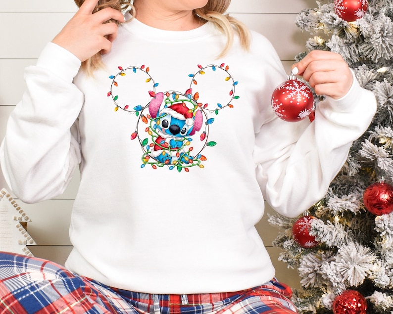 Może przedstawiać: Biała bluza z niebieską postacią Stitch, kt&oacute;ra ma na sobie czapkę Świętego Mikołaja i jest zaplątana w świąteczne lampki. Lampki mają kształt uszu Myszki Miki.