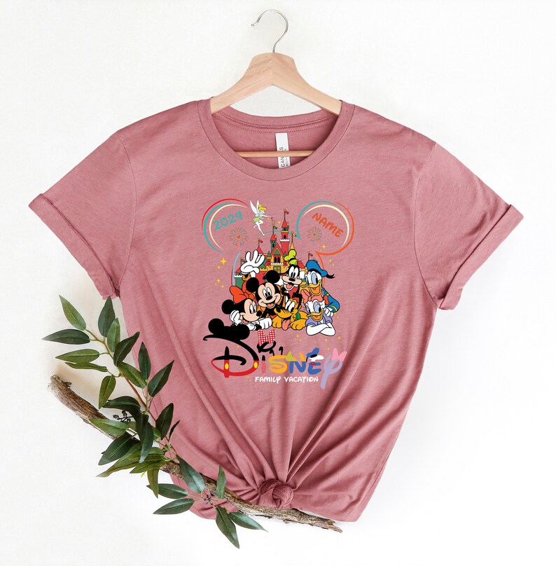 Puede incluir: Una camiseta rosa con un gr&aacute;fico de Mickey Mouse y sus amigos frente al Castillo de Cenicienta. El texto "Disney Family Vacation" est&aacute; debajo del gr&aacute;fico. El a&ntilde;o "2024" y la palabra "NAME" est&aacute;n en burbujas de di&aacute;logo sobre el castillo.