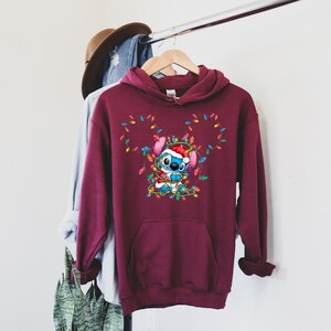 Może przedstawiać: Bordowa bluza z kapturem z grafiką świąteczną przedstawiającą Stitch'a z Lilo & Stitch'a zaplątanego w świąteczne lampki. Stitch ma na sobie czapkę Świętego Mikołaja.