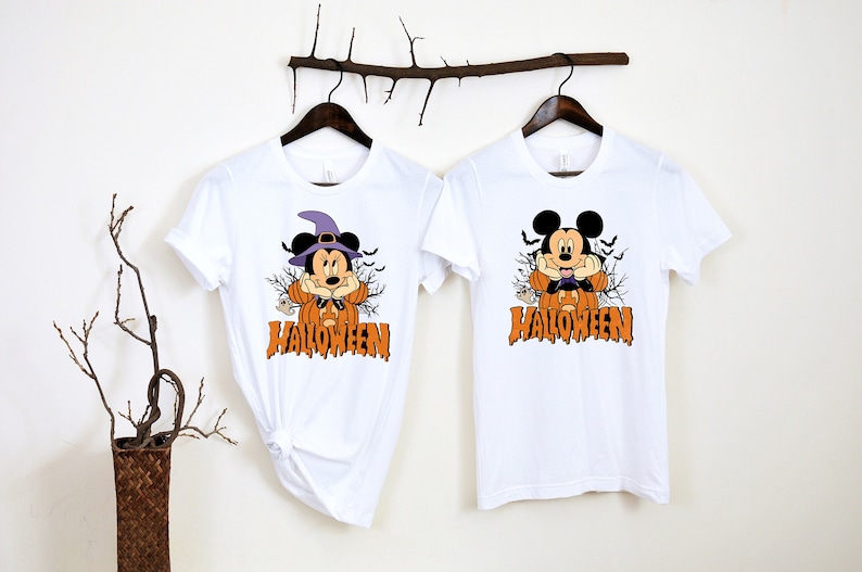 K&ouml;nnte beinhalten: Zwei wei&szlig;e T-Shirts mit Halloween-Motiv. Ein Shirt zeigt Mickey Mouse als Hexe mit dem Text "HALLOWEEN". Das andere Shirt zeigt Mickey Mouse als K&uuml;rbis mit dem Text "HALLOWEEN".