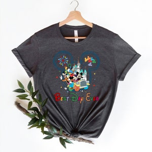 May include: A dark gray t-shirt with a colorful graphic of Mickey Mouse and friends riding in a car in front of the Cinderella Castle. The graphic is inside a blue Mickey Mouse ear silhouette. The text "Best Day Ever" is printed below the graphic in rainbow colors.