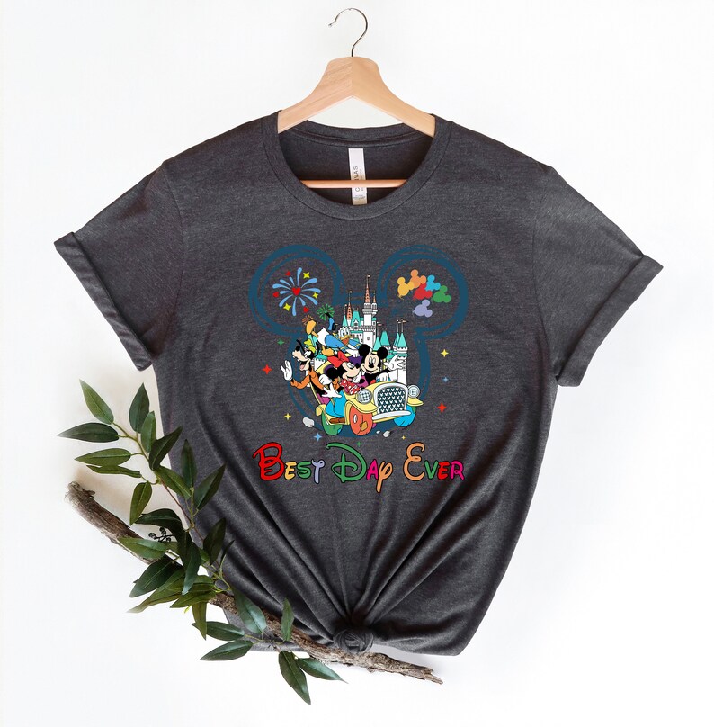 May include: A dark gray t-shirt with a colorful graphic of Mickey Mouse and friends riding in a car in front of the Cinderella Castle. The graphic is inside a blue Mickey Mouse ear silhouette. The text "Best Day Ever" is printed below the graphic in rainbow colors.