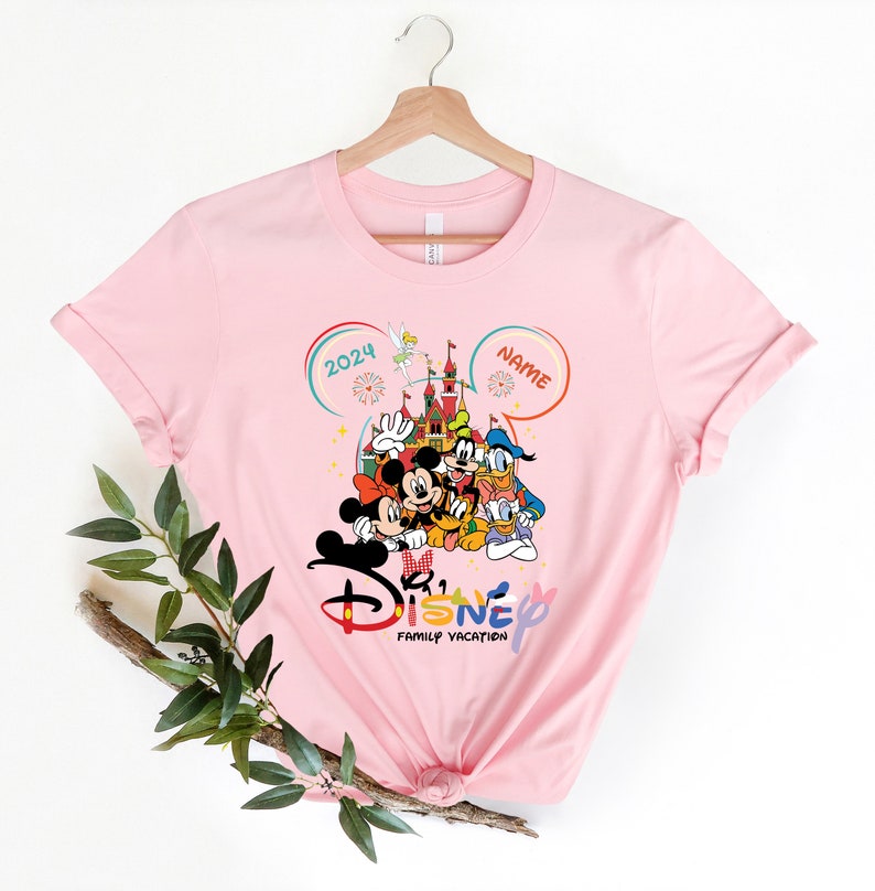 Puede incluir: Camiseta rosa con un gr&aacute;fico de Mickey Mouse y sus amigos frente al Castillo de Cenicienta. El texto "2024" y "NAME" est&aacute;n en las esquinas superiores del gr&aacute;fico. El texto "Disney Family Vacation" est&aacute; debajo del gr&aacute;fico.