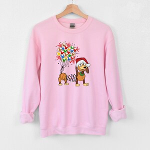 Pu&ograve; includere: Felpa rosa chiaro con un cane dei cartoni animati con un cappello da Babbo Natale e palloncini. I palloncini presentano facce di personaggi. Il cane ha un corpo a molla. Un design festivo per le vacanze.