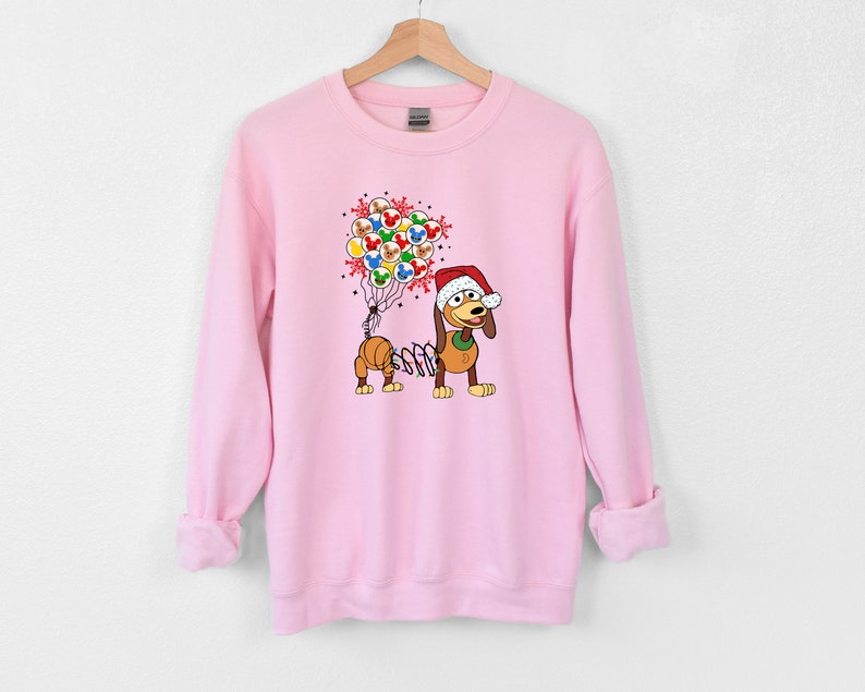 Pu&ograve; includere: Felpa rosa chiaro con un cane dei cartoni animati con un cappello da Babbo Natale e palloncini. I palloncini presentano facce di personaggi. Il cane ha un corpo a molla. Un design festivo per le vacanze.