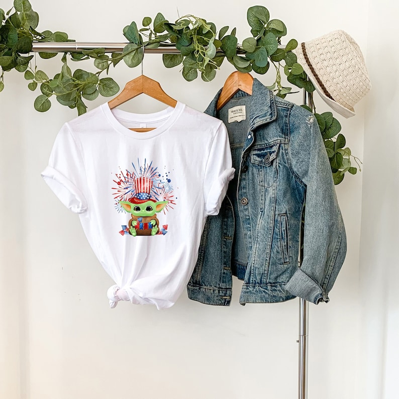 May include: White t-shirt with a green character wearing a red, white, and blue hat and holding a red, white, and blue flag. The character is surrounded by red, white, and blue fireworks. The t-shirt is on a hanger with a denim jacket next to it.