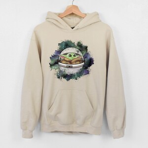 May include: A beige hoodie with a graphic print of a green alien character in a spaceship against a galaxy background.