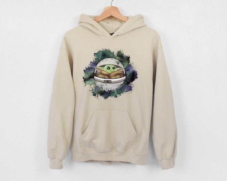May include: A beige hoodie with a graphic print of a green alien character in a spaceship against a galaxy background.