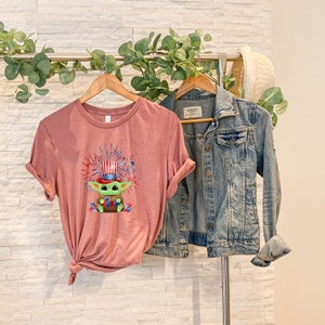 May include: A mauve t-shirt with a green character wearing a red, white, and blue hat and holding a firework. The character is surrounded by fireworks. The t-shirt is hanging on a rack with a denim jacket.