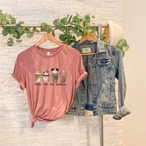 May include: A pink t-shirt with a graphic of four different coffee cups with Mickey Mouse ears and the text "Here for the drinks".