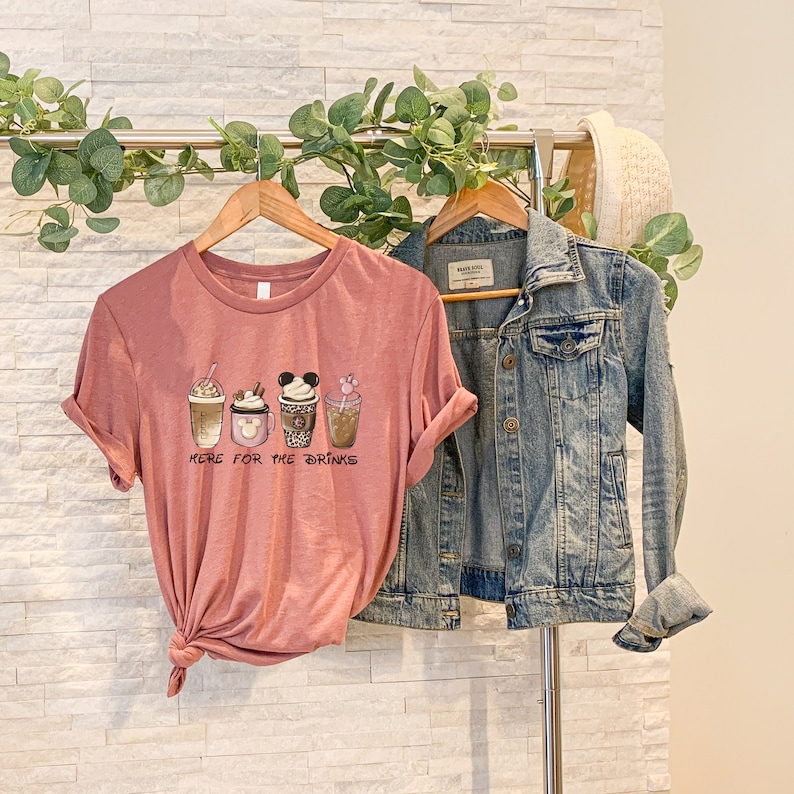 May include: A pink t-shirt with a graphic of four different coffee cups with Mickey Mouse ears and the text "Here for the drinks".