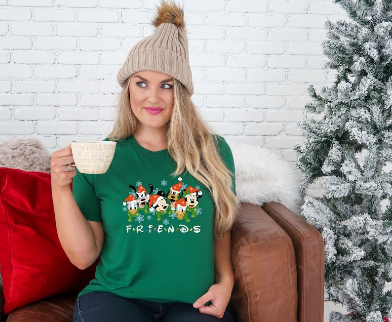 May include: A green t-shirt with a Christmas-themed graphic of Mickey Mouse and his friends. The graphic features the characters wearing Santa hats and surrounded by snowflakes. The text "FRIENDS" is printed below the graphic.