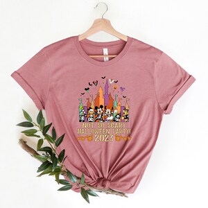 May include: A pink t-shirt featuring a graphic of Mickey Mouse and his friends in front of a castle with the text "Not So Scary Halloween Party! 2023"