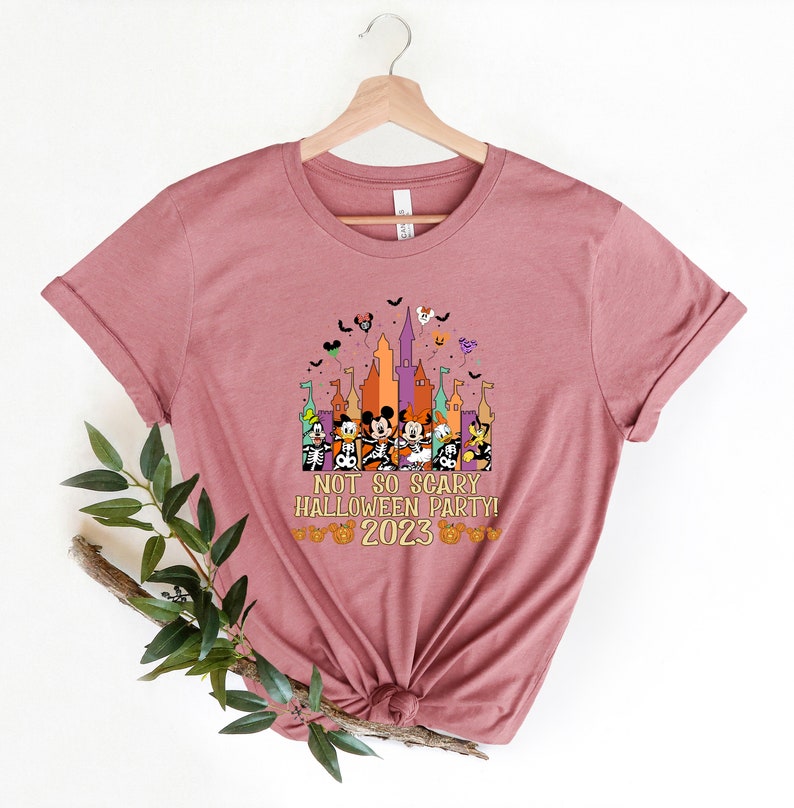 May include: A pink t-shirt featuring a graphic of Mickey Mouse and his friends in front of a castle with the text "Not So Scary Halloween Party! 2023"