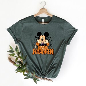 K&ouml;nnte beinhalten: Ein dunkelgr&uuml;nes T-Shirt mit einem Cartoon-Bild von Mickey Mouse, der als Vampir verkleidet ist. Mickey h&auml;lt eine K&uuml;rbislaterne und hinter ihm ist ein Geist. Der Text "HALLOWEEN" ist unter Mickey in tropfenden orangefarbenen Buchstaben gedruckt.