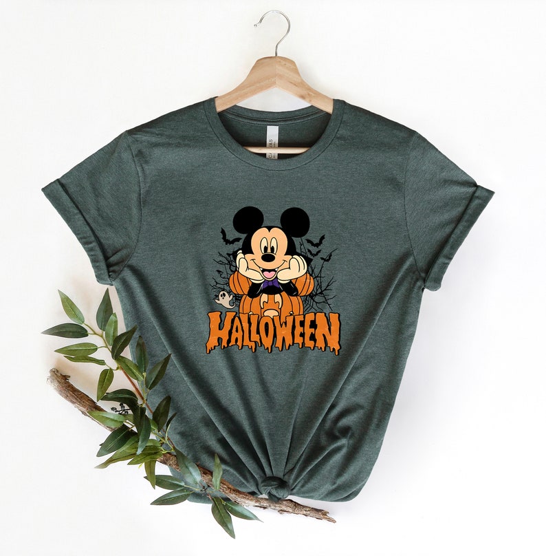 K&ouml;nnte beinhalten: Ein dunkelgr&uuml;nes T-Shirt mit einem Cartoon-Bild von Mickey Mouse, der als Vampir verkleidet ist. Mickey h&auml;lt eine K&uuml;rbislaterne und hinter ihm ist ein Geist. Der Text "HALLOWEEN" ist unter Mickey in tropfenden orangefarbenen Buchstaben gedruckt.