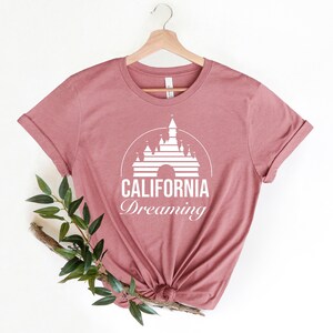 Puede incluir: Una camiseta color malva con un gr&aacute;fico blanco del castillo de la Bella Durmiente y el texto "CALIFORNIA Dreaming".