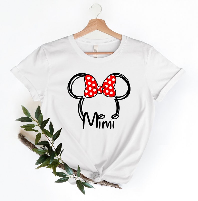 Op de afbeelding: Wit T-shirt met een zwart-witte omtrek van Mickey Mouse oren met een rode strik met witte stippen. De tekst "Mimi" staat onder de oren.