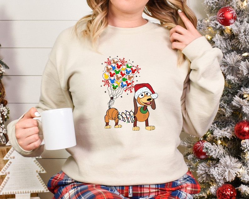 Pu&ograve; includere: Felpa color crema con un bassotto dei cartoni animati con un cappello da Babbo Natale e luci natalizie. Il bassotto tiene in mano dei palloncini di Topolino. Abbigliamento festivo per le vacanze.