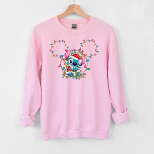 Może przedstawiać: R&oacute;żowa bluza z niebieską postacią Stitch, kt&oacute;ra ma czapkę Świętego Mikołaja i trzyma świąteczne lampki w kształcie uszu Myszki Miki.