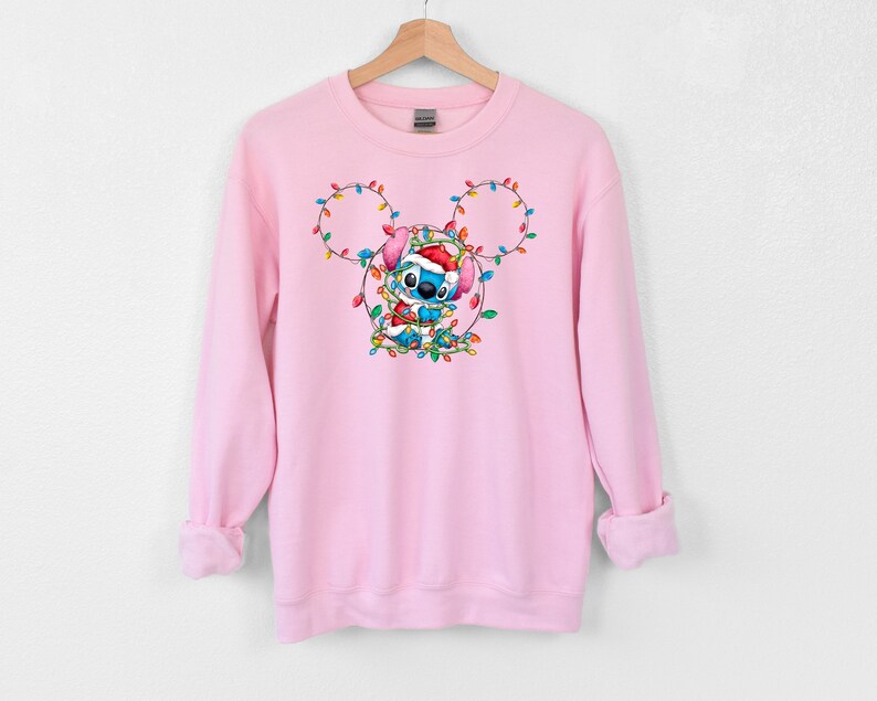 Może przedstawiać: R&oacute;żowa bluza z niebieską postacią Stitch, kt&oacute;ra ma czapkę Świętego Mikołaja i trzyma świąteczne lampki w kształcie uszu Myszki Miki.