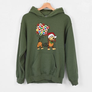 Pu&ograve; includere: Una felpa con cappuccio verde oliva con un bassotto dei cartoni animati che indossa un cappello da Babbo Natale e tiene in mano dei palloncini. Il cane ha un corpo a molla e tiene in mano un gruppo di palloncini colorati. La felpa ha una tasca frontale e un cappuccio.
