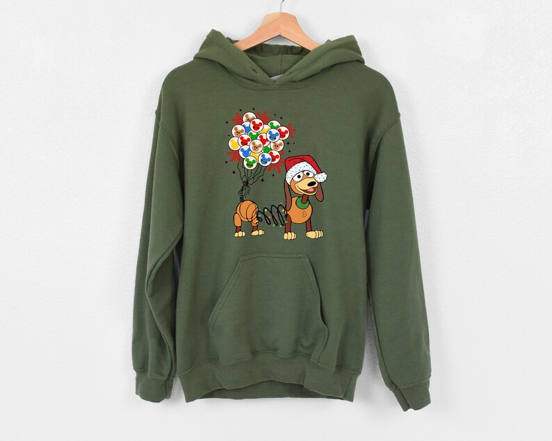 Pu&ograve; includere: Una felpa con cappuccio verde oliva con un bassotto dei cartoni animati che indossa un cappello da Babbo Natale e tiene in mano dei palloncini. Il cane ha un corpo a molla e tiene in mano un gruppo di palloncini colorati. La felpa ha una tasca frontale e un cappuccio.