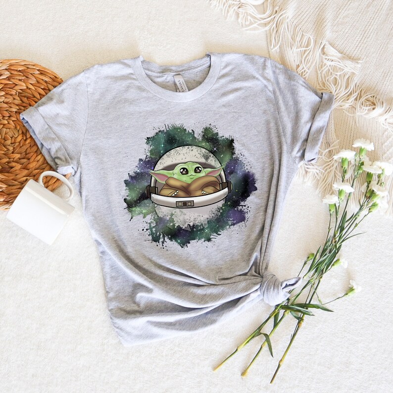 May include: A gray t-shirt with a graphic of a green alien character in a space helmet against a galaxy background.