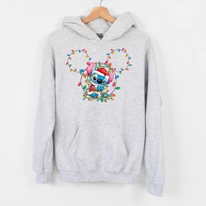 Może przedstawiać: Szara bluza z kapturem z niebieską postacią Stitch, kt&oacute;ra nosi czapkę Świętego Mikołaja i jest otoczona kolorowymi świątecznymi światełkami w kształcie uszu Myszki Miki.