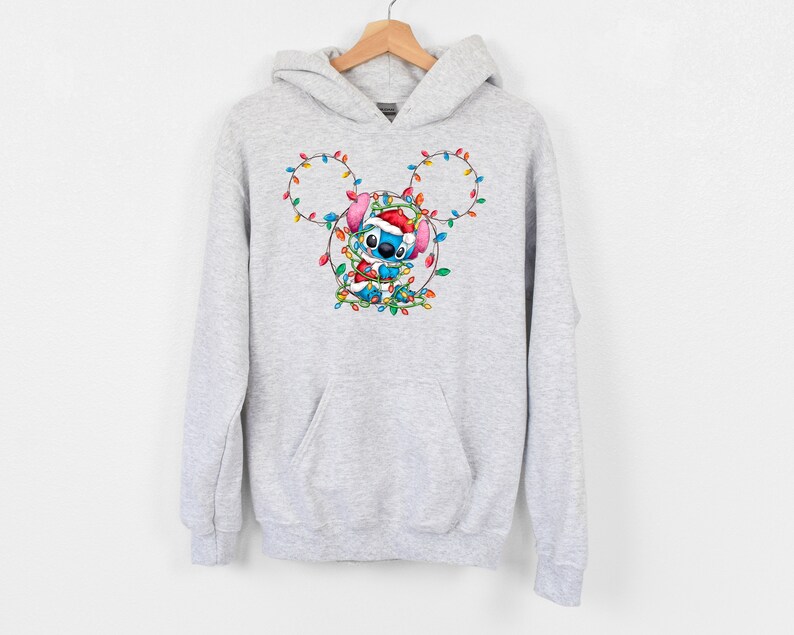 Może przedstawiać: Szara bluza z kapturem z niebieską postacią Stitch, kt&oacute;ra nosi czapkę Świętego Mikołaja i jest otoczona kolorowymi świątecznymi światełkami w kształcie uszu Myszki Miki.