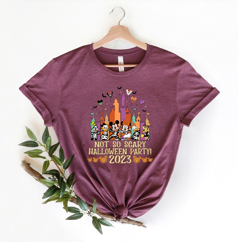 May include: A maroon t-shirt featuring a graphic of Mickey Mouse and his friends in front of a castle. The text "Not so scary Halloween party! 2023" is printed on the shirt.