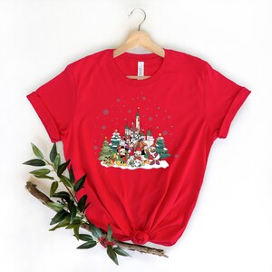 May include: Red t-shirt with a Christmas-themed graphic featuring Mickey Mouse and friends in front of a snow-covered castle. The graphic also includes Christmas trees and snowflakes.