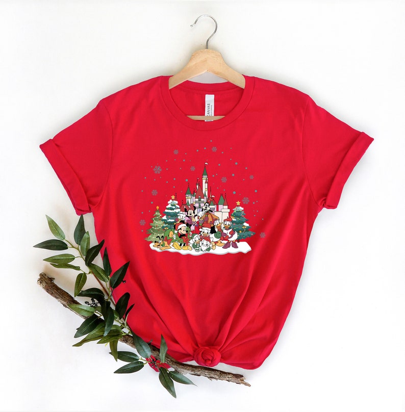 May include: Red t-shirt with a Christmas-themed graphic featuring Mickey Mouse and friends in front of a snow-covered castle. The graphic also includes Christmas trees and snowflakes.