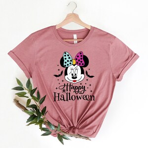 Puede incluir: Una camiseta color malva con un gr&aacute;fico de un rat&oacute;n de dibujos animados con un lazo rosa y azul, murci&eacute;lagos y estrellas. El texto "Happy Halloween" est&aacute; impreso debajo del rat&oacute;n.