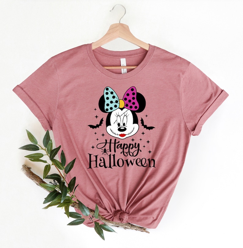 Puede incluir: Una camiseta color malva con un gr&aacute;fico de un rat&oacute;n de dibujos animados con un lazo rosa y azul, murci&eacute;lagos y estrellas. El texto "Happy Halloween" est&aacute; impreso debajo del rat&oacute;n.