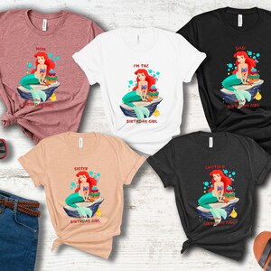 K&ouml;nnte beinhalten: Eine Auswahl an T-Shirts in verschiedenen Farben, jeweils mit einem Cartoon-Meerjungfrau-Design. Auf den Shirts steht "Mama", "Ich bin das Geburtstagskind", "Papa", "Schwester" und "Bruder". Die Meerjungfrau hat rotes Haar und einen t&uuml;rkisfarbenen Schwanz.