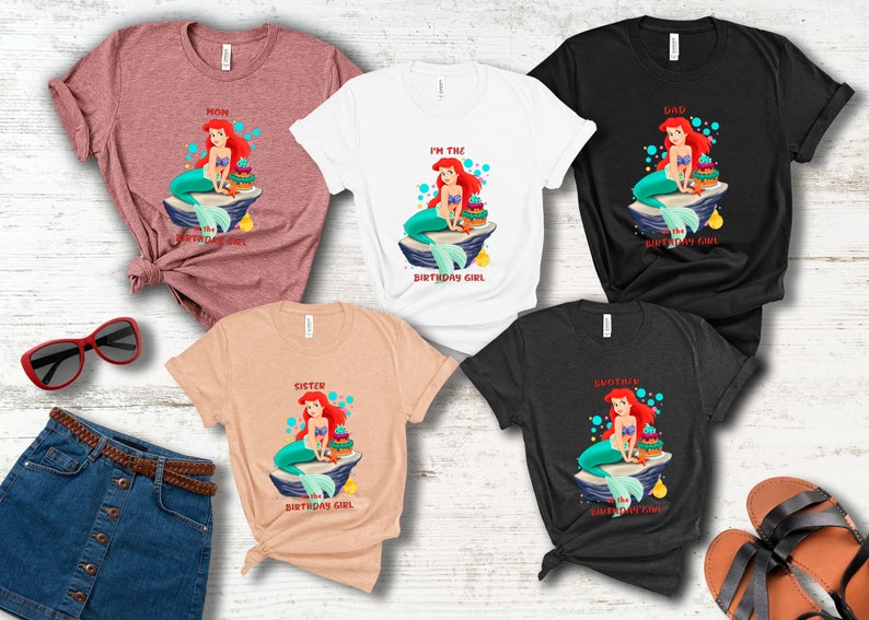 K&ouml;nnte beinhalten: Eine Auswahl an T-Shirts in verschiedenen Farben, jeweils mit einem Cartoon-Meerjungfrau-Design. Auf den Shirts steht "Mama", "Ich bin das Geburtstagskind", "Papa", "Schwester" und "Bruder". Die Meerjungfrau hat rotes Haar und einen t&uuml;rkisfarbenen Schwanz.