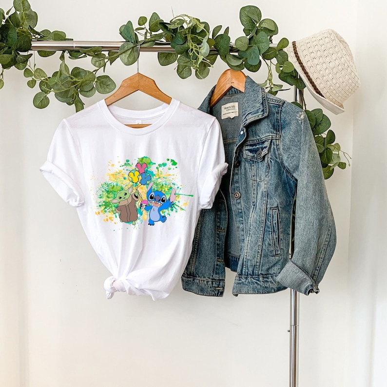 K&ouml;nnte beinhalten: Wei&szlig;es T-Shirt mit einer bunten Grafik von Stitch und Baby Yoda, die Ballons halten. Das Shirt h&auml;ngt an einem Kleiderb&uuml;gel mit einer Jeansjacke und einem Strohhut.