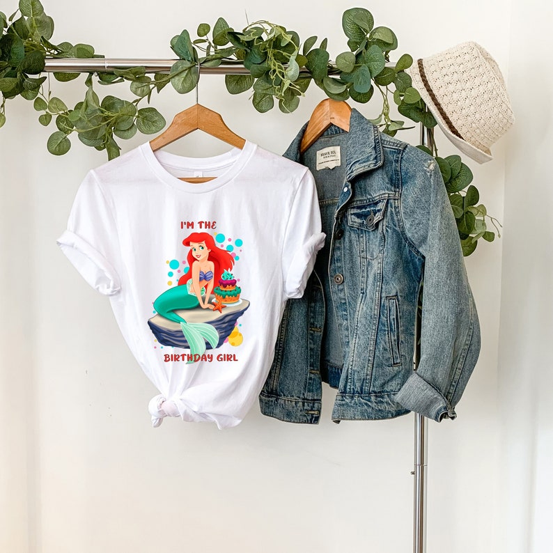 K&ouml;nnte beinhalten: Wei&szlig;es T-Shirt mit einem farbenfrohen Aufdruck einer Meerjungfrau mit roten Haaren und einem t&uuml;rkisfarbenen Schwanz, die auf einem Felsen mit Geburtstagskuchen sitzt. Der Text "I'M THE BIRTHDAY GIRL" ist darunter gedruckt. Eine Jeansjacke und ein Strohhut sind ebenfalls zu sehen.