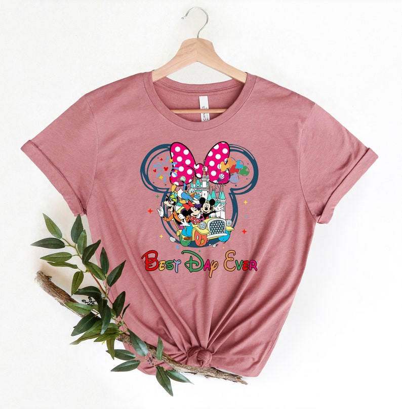 May include: A pink t-shirt with a graphic of Mickey Mouse and friends in a car in front of the Cinderella Castle. The graphic is inside a large Mickey Mouse ear with a pink bow. The text "Best Day Ever" is below the graphic.