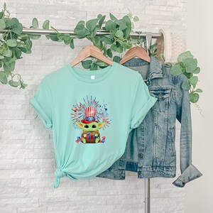 May include: A light blue t-shirt with a graphic of a green character wearing a red, white, and blue hat and holding fireworks. The character is surrounded by red, white, and blue fireworks.