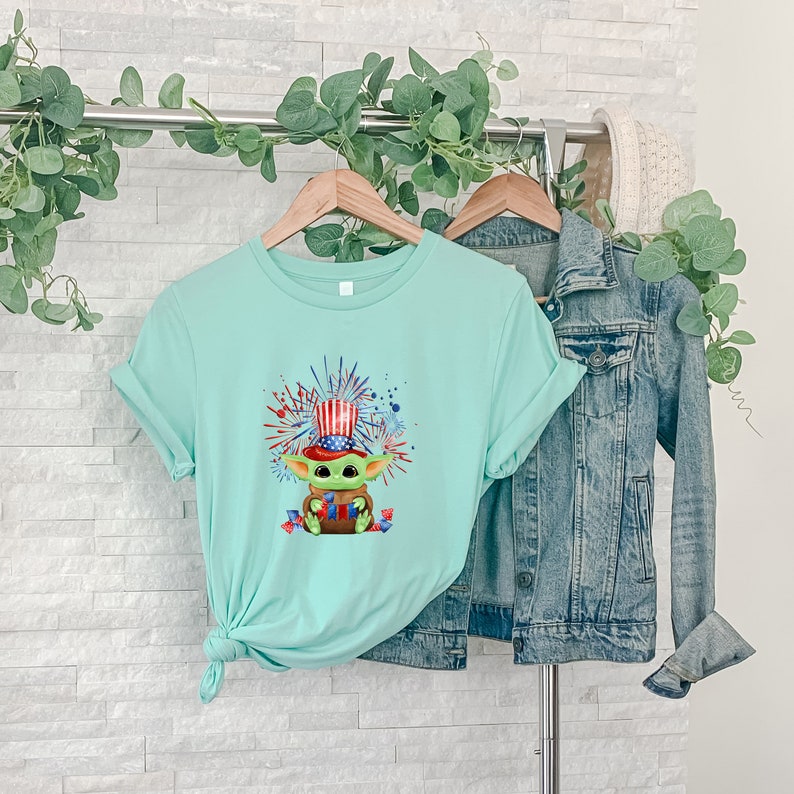 May include: A light blue t-shirt with a graphic of a green character wearing a red, white, and blue hat and holding fireworks. The character is surrounded by red, white, and blue fireworks.