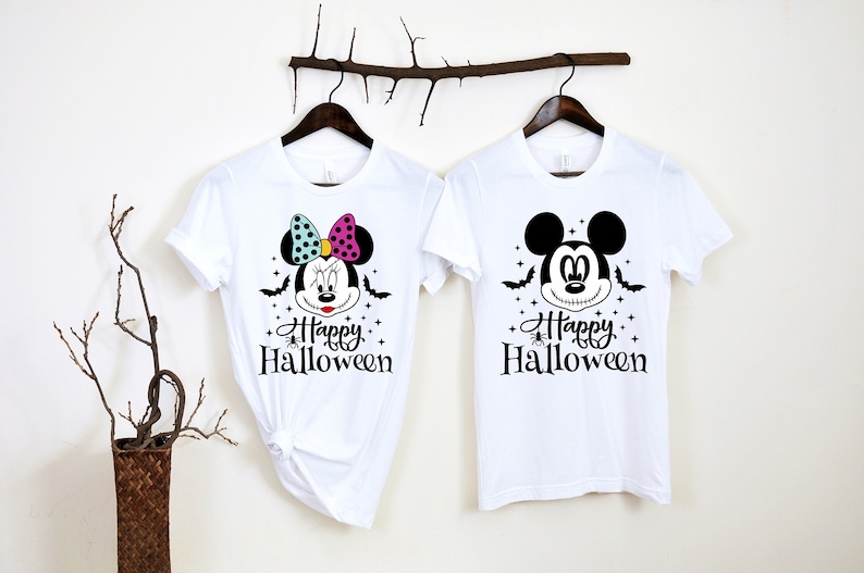 Puede incluir: Dos camisetas blancas con tem&aacute;tica de Halloween. Una camiseta presenta un Mickey Mouse en blanco y negro con el texto "Happy Halloween". La otra camiseta presenta una Minnie Mouse colorida con el texto "Happy Halloween".